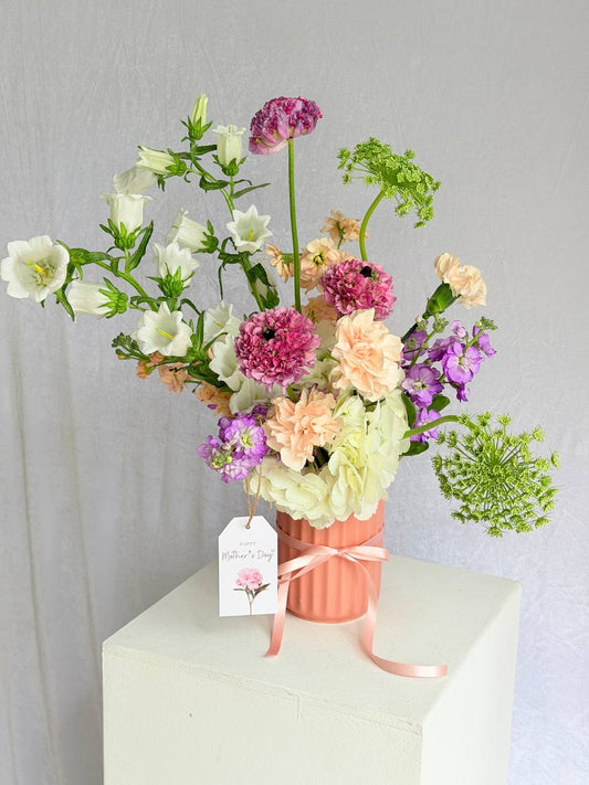 Bouquet of flowers in a pink vase with a 'Mother's Day' tag on a white surface.