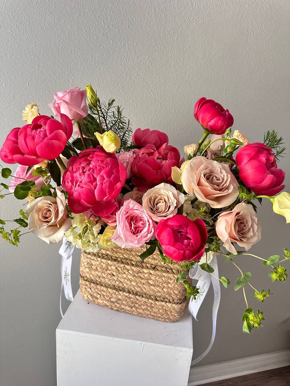 Bouquet of pink and beige flowers in a woven basket on a white surface with a gray wall background.