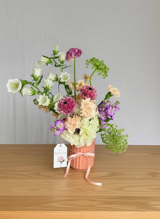 Bouquet of flowers in a pink vase with a 'Mother's Day' tag on a white surface.