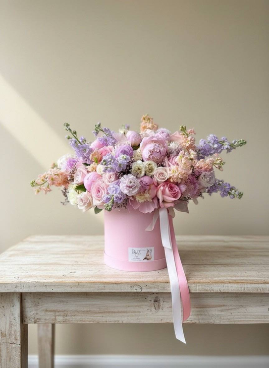Pink flower box with a bouquet of flowers on a wooden table against a beige wall.
