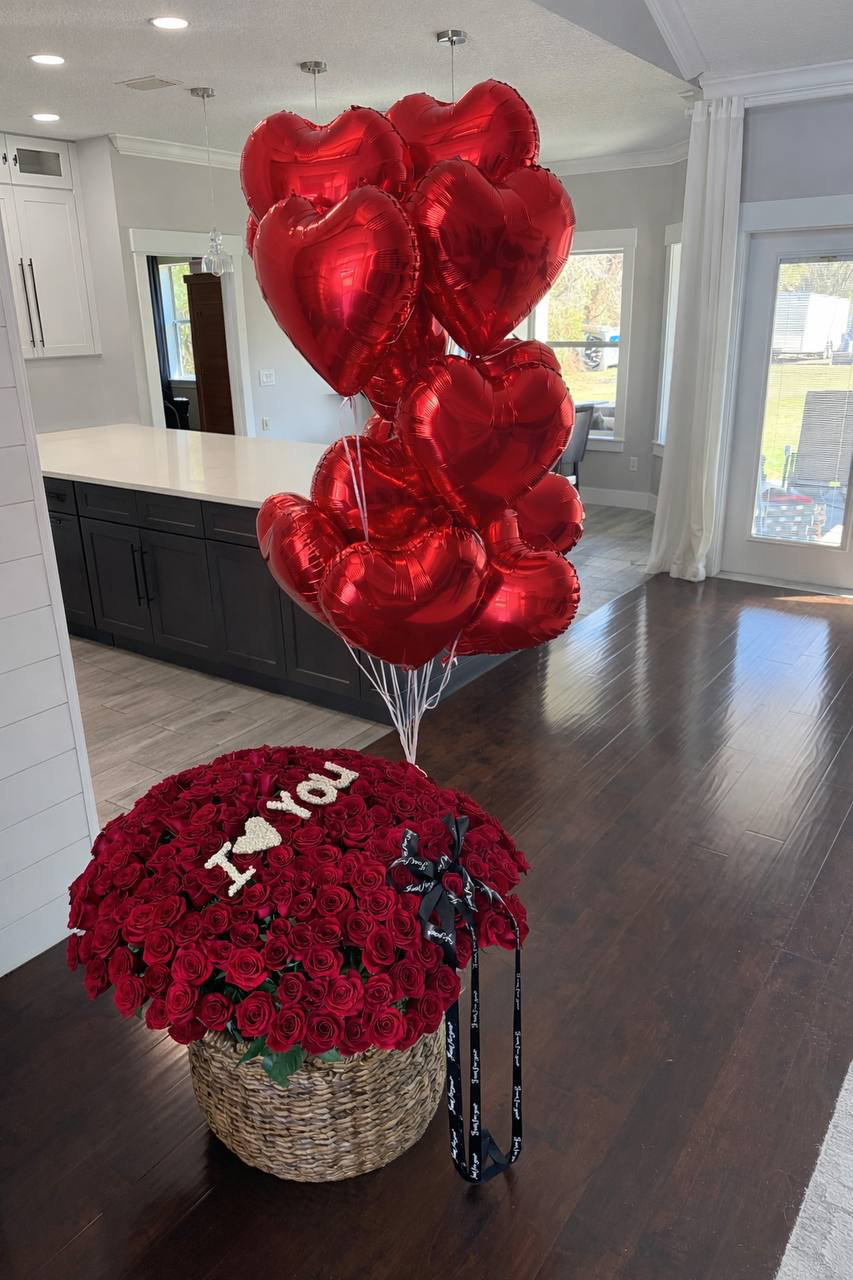 Red heart-shaped balloons and a bouquet of red roses on a wooden floor in a home setting.